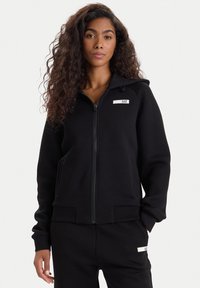 Sudadera negra con cremallera y capucha, confeccionada en tela suave. Presenta un pequeño logo blanco, bolsillos frontales y puños y dobladillo de canalé.