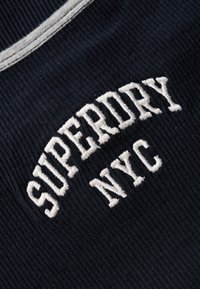 Navyblauer Stoff mit gerippter Textur, der weißen gestickten Text "SUPERDRY NYC" in einem Bogen zeigt, mit kontrastierenden Nähdetails.