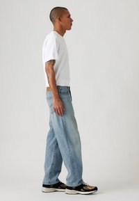 Witte korte mouwen t-shirt, lichtblauwe losse jeans en zwarte sneakers met tan accenten. Zicht van opzij toont een ontspannen stijl en pasvorm.