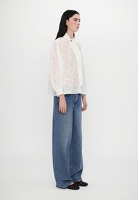 Blusa bianca trasparente con motivo floreale, maniche arricciate e colletto con bottoni, abbinata a jeans in denim a gamba ampia e scarpe nere a pois.