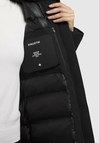 Crna puffer jakna s glatkom površinom, s logom na zakrpi i zip džepom na unutarnjoj podstavi. Mekana tekstura i izolirani dizajn.