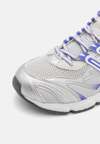 Silver löparsko med mesh-övre, lila detaljer och texturerad sula. Har intrikata sömmar och snörhål för en säker passform.