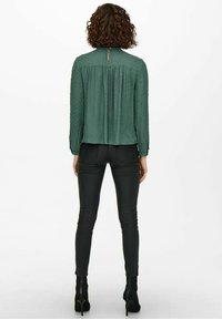 Dunkelgrüne, strukturierte Bluse mit plisseeartigem Rücken und Knopfverschluss, kombiniert mit taillierten schwarzen Lederhosen und schwarzen High-Heels.