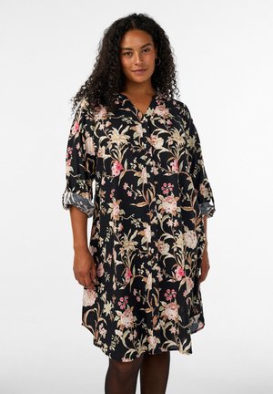 Vrouw met krullend haar die een zwarte, knielange bloemenjurk met opgerolde mouwen draagt tegen een effen witte achtergrond.
