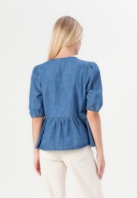 Denim peplum top s krátkymi puffovými rukávmi, okrúhlym výstrihom a gathered elastickým pásom. Svetlo modrá farba s hladkým povrchom.