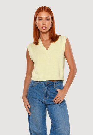 Giovane donna con capelli rossi che indossa un top giallo pallido senza maniche con scollo a V e jeans blu a vita alta, in piedi davanti a uno sfondo semplice.