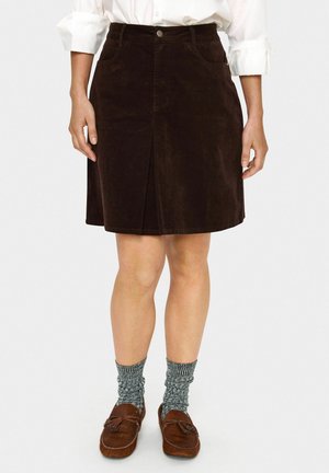 Brauner Cordrock mit vorderem Schlitz, zwei seitlichen Taschen und Knopfverschluss. Getragen mit grauen gestreiften Socken und braunen Mokassins.
