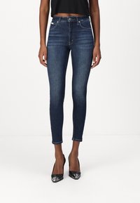 Jeans skinnies de color azul oscuro con una cintura alta, presentan un diseño clásico de cinco bolsillos y un ligero desvanecimiento, combinados con tacones negros de punta afilada.