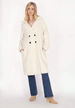 Cappotto doppio petto color crema, soffice, con bottoni neri, tasche laterali e colletto a revers, indossato sopra jeans a zampa d'elefante in denim.