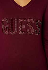 Burgunderfarbener Stricktop mit V-Ausschnitt und großem 'GUESS'-Logo in strukturierten, metallischen Akzenten entlang der Buchstaben. Glatte, enganliegende Stoffqualität.