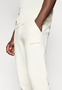 Cremefarvede joggers i blødt stof med sidelommer og en subtil trykt logo på låret. Glat tekstur, tætsiddende design.
