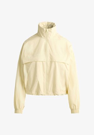 Világos bézs, cipzáras szélálló dzseki magas gallérral, rugalmas mandzsettákkal és a mellkason és a háton végigfutó húzóvarrással.