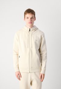Ung mand iført en cremefarvet zip-up hættetrøje og matchende bukser, stående foran en ensfarvet lys grå baggrund.