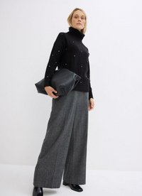 Maglione nero a collo alto con piccole decorazioni, abbinato a pantaloni grigi a gamba larga e che tiene una pochette di pelle nera.