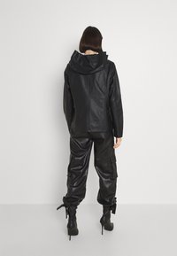 Veste noire en simili cuir avec capuche, associée à un pantalon noir en simili cuir ample, comportant des poches cargo et des détails à lacets au niveau des chevilles.