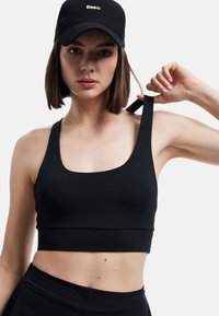Sort sportstop med rund udskæring og brede stropper; blødt, strækbart materiale; minimalistisk design. Modellen bærer en sort kasket med hvid tekst.