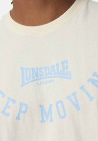 Kreminė medvilninė marškinė su šviesiai mėlyna spauda, "LONSDALE LONDON" ir "KEEP MOVING", su puraus apykakle.