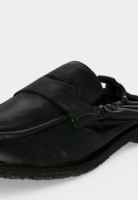Mocassin en cuir noir avec arrière élastiqué et semelle texturée, montré sous un léger angle latéral sur fond blanc.