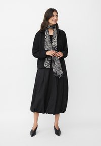 Giacca nera e strutturata sopra un vestito nero e voluminoso. Una sciarpa a motivi bianco e nero aggiunge un tocco di dettaglio. Scarpe nere a punta completano il look.