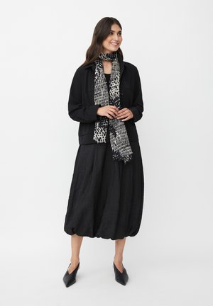 Zwarte, gestructureerde jacket over een zwarte, volumineuze jurk. Een zwart-witte, patronen sjaal voegt detail toe. Puntige zwarte schoenen maken de look compleet.