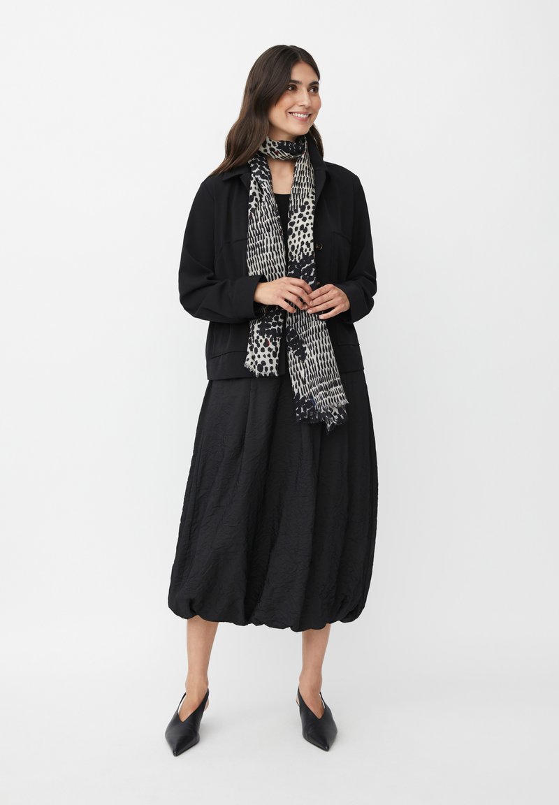 Giacca nera e strutturata sopra un vestito nero e voluminoso. Una sciarpa a motivi bianco e nero aggiunge un tocco di dettaglio. Scarpe nere a punta completano il look.
