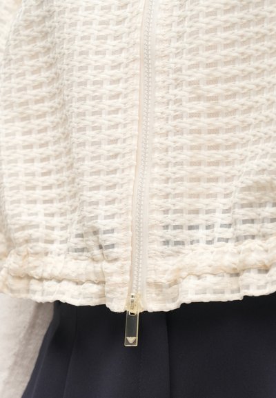 Λευκό μπουφάν με φερμουάρ από υφαντό ύφασμα με υφή, με διαφανές τμήμα στο φερμουάρ και σκοτεινό ύφασμα από κάτω.