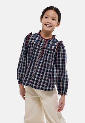 Fille souriante portant une blouse à carreaux bleu marine avec bordure rouge et broderie florale, associée à un pantalon large beige clair sur fond blanc.