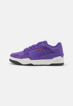 Baskets basses violettes avec panneaux en daim et tissu, semelle intermédiaire blanche, semelle extérieure noire et étiquette orange "GNAP !" sur le côté avec le logo Puma.