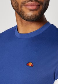 Blaues Baumwoll-T-Shirt mit rundem Ausschnitt und einem kleinen bestickten Logo in Rot, Gelb und Orange auf der linken Seite. Weiche Textur.