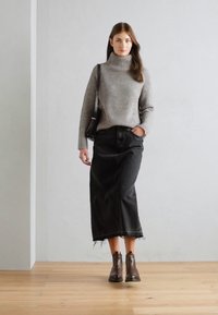 Pull gris à côtes, jupe en denim noire avec ourlet effiloché, et bottines marron. Le modèle se tient sur un sol en bois contre un mur clair.