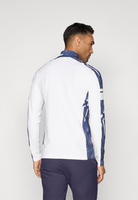 Chemise de sport à manches longues en blanc, avec des accents en bleu marine à motifs sur les épaules et les manches, dotée d'un col montant et d'une coupe ajustée.