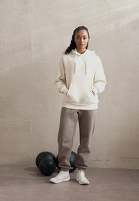 Felpa leggera color crema con tasca a marsupio, abbinata a pantaloni della tuta grigi. Presenta cordini e polsini a costine. Sneakers bianche e palloni neri sullo sfondo.