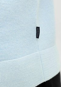 Pull bleu clair en tricot avec ourlet côtelé et fente latérale. Les étiquettes sont visibles avec des inscriptions noires pour l'identification de la marque.