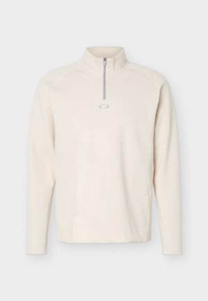 Pull à manches longues crème avec une demi-fermeture éclair à l'avant et un petit logo ovale gris sous la fermeture éclair sur la poitrine.