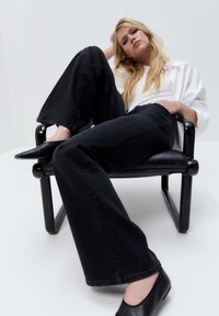 Jeans neri a gamba larga in denim, abbinati a una camicia bianca con bottoni. Modello seduto su una sedia moderna nera, che mostra uno stile casual.