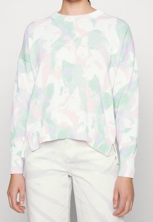 Personne portant un pull à motif camouflage pastel en rose clair, vert et violet avec un pantalon blanc, visible de la poitrine jusqu'aux hanches.