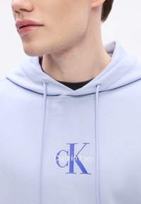 Svetlomodrá mikina s jemnou textúrou, vybavená bielym logom "Calvin Klein" a fialovým dizajnom "CK". Na golieri sú viditeľné zťahovacie šnúrky.