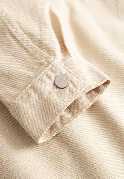 Cream-colored fabric sleeve with visible stitching and a round silver snap button featuring an engraved logo.
