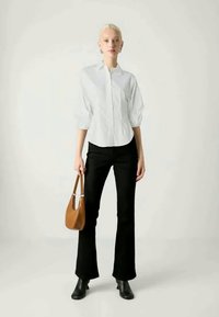 Vrouw in een witte button-up blouse en zwarte broek, met een bruine handtas, staande tegen een effen lichte achtergrond.