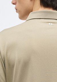 Κοντινό πλάνο στο άνω μέρος της πλάτης και του λαιμού ενός ατόμου που φοράει μπεζ πουκάμισο με γιακά και ένα μικρό λευκό λογότυπο κοντά στον γιακά.