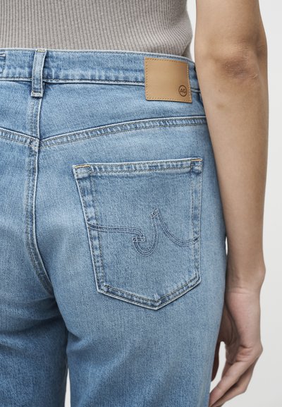 Ανοιχτό μπλε τζιν με τσέπη πίσω που έχει κεντημένο σχέδιο, φορεμένα από άτομο με γκρι ριμπ τοπ, δείχνοντας μερικό χέρι και μπράτσο.