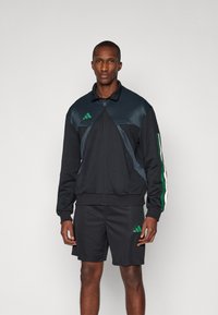Veste de sport zippée noire avec des rayures vertes et blanches, en matériau texturé et au design épuré ; associée à un short en mesh noir.