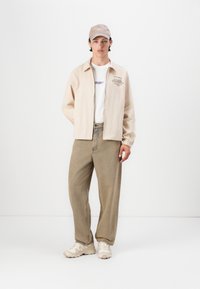 Jachetă crem cu fermoar, cu manșete elastice, purtată peste un tricou alb cu text violet, asortată cu pantaloni largi crem și adidași de culoare deschisă.