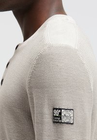 Beige gestreiftes Langarmshirt mit Knopfverschluss und Rundhalsausschnitt. Besitzt ein schwarzes Logopatch am Ärmel. Strukturierter Stoff.