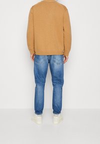 Suéter de punto beige con bordes acanalados, combinado con jeans azul claro y zapatillas blancas. Costuras visibles y detalles de bolsillo en los jeans.