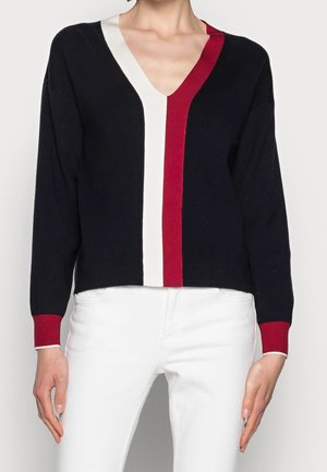Femme portant un pull noir à col en V avec des rayures verticales blanches et rouges et des poignets rouges, associé à un pantalon blanc.