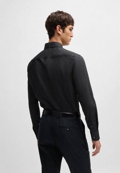 Chemise noire à manches longues, col classique et texture lisse, associée à un pantalon foncé à motifs et une ceinture noire.