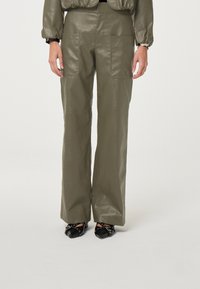Olijfgroene neplederen wijde broek met twee voorzakken, een soepele textuur en een hoge taille. Zwarte schoenen met decoratieve accenten.