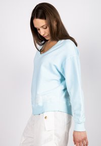 Light blue oversized sweatshirt with a V-neckline and long sleeves. Features white embroidered text at the hem. Soft fabric texture.
