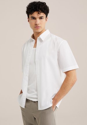 Noor mees lokkis juustega, kellel on peas valge lühikeste varrukatega avatud särk valge maika ja beežide pükstega, käed taskutes, neutraalne taust.